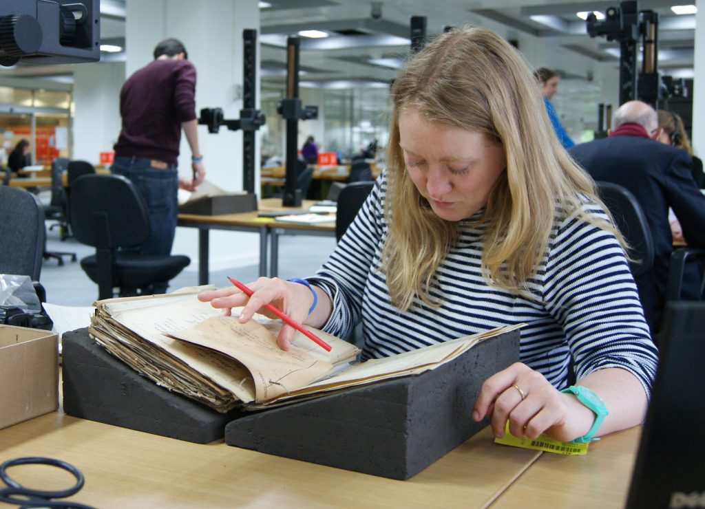 A color photograph of a researcher seated at a table in the archives reading room, looking at a manuscript that is resting in a foam book cradle. In the background is another researcher using a digital camera to photograph archival material.