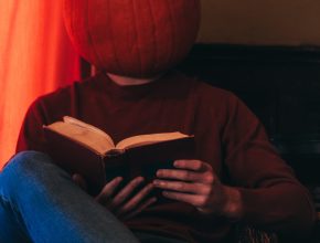 Photograph of a person sitting in a chair with an open book propped on a crossed leg. The person's head is a pumpkin.