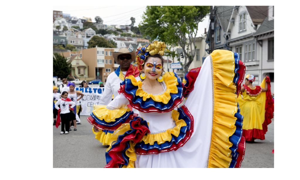 Woman in colorful dress with her face painted smiles into the camera while marching in a parade.