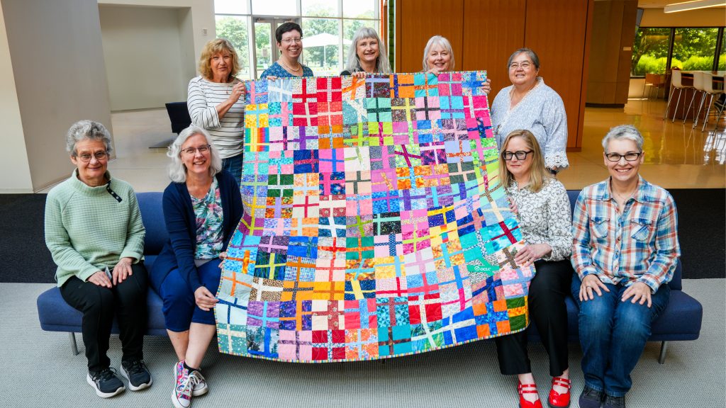 Nine people in a bright lobby pose with a large, colorful patchwork quilt featuring a grid of multicolored fabric squares; four stand behind holding the quilt Four people sit on a blue bench in front and five stand behind the bench