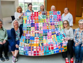 Nine people in a bright lobby pose with a large, colorful patchwork quilt featuring a grid of multicolored fabric squares; four stand behind holding the quilt Four people sit on a blue bench in front and five stand behind the bench